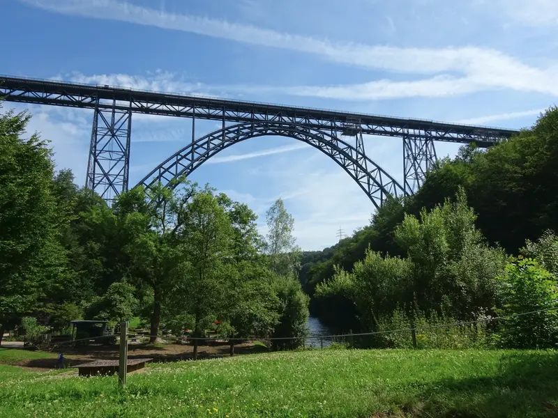 Im Vordergrund eine grüne Wiese und mittig der Fluss "Wupper". Beidseits davon steigen bewaldete Hänge empor, deren Bäume in vollen Grün stehen und über denen ein hellblauer Himmel mit weißen Schleierwolken zu sehen ist. Die Bildmitte zeigt eine filigran wirkende, aus dünnen, schwarzen Stahlstreben konstruierte, hohe Eisenbahnbrücke, welche den Fluss und das Tal überspannt. Das Doppelgleis wird von einem Stahl-Fachwerkbogen und mehreren Stahl-Fachwerksäulen tragen, durch die der dahinter liegende Himmel erkennbar ist.