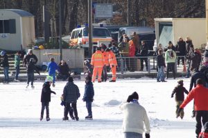 Erwachsene und Kinder in Winterbekleidung laufen auf dem zugefrorenen Uelfebad Schlittschuh. Drei Angehörige der DLRG beaufsichtigen das bunte Treiben