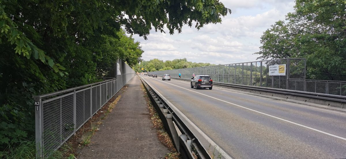 Zwischen hohen Schutzzäunen führt die Straße über die Blombacher Bach Brücke. Mehrere Autos und Radfahrer fahren gerade über die Brücke.