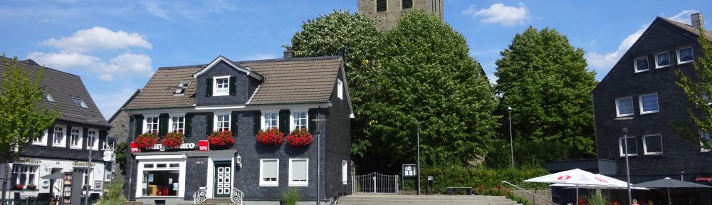 Neben dem Radevormwalder Marktplatz steht hinter Bäumen der Kirchturm der evangelisch-reformierten Kirche mit geschieferter Zwiebelspitze und Turmuhr