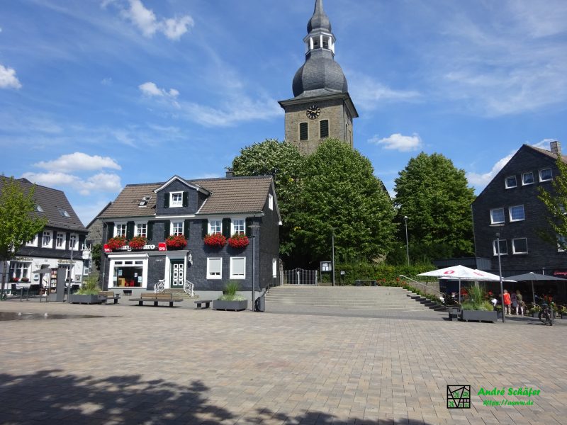 Neben dem Radevormwalder Marktplatz steht hinter Bäumen der Kirchturm der evangelisch-reformierten Kirche mit geschieferter Zwiebelspitze und Turmuhr