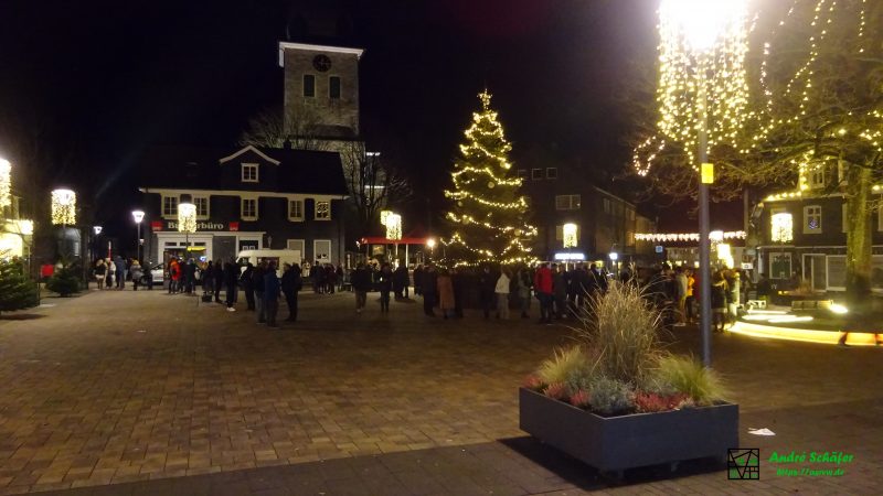 Auf dem weihnachtlich geschmückten Marktplatz stehen um Mitternacht viele Menschen und hören das Geläut der Kirche zum 1. Weihnachtstag