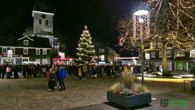 Zahlreiche Menschen haben sich für Mitternacht auf dem Marktplatz um einen beleuchteten Weihnachtsbaum versammelt und singen Weihnachtslieder